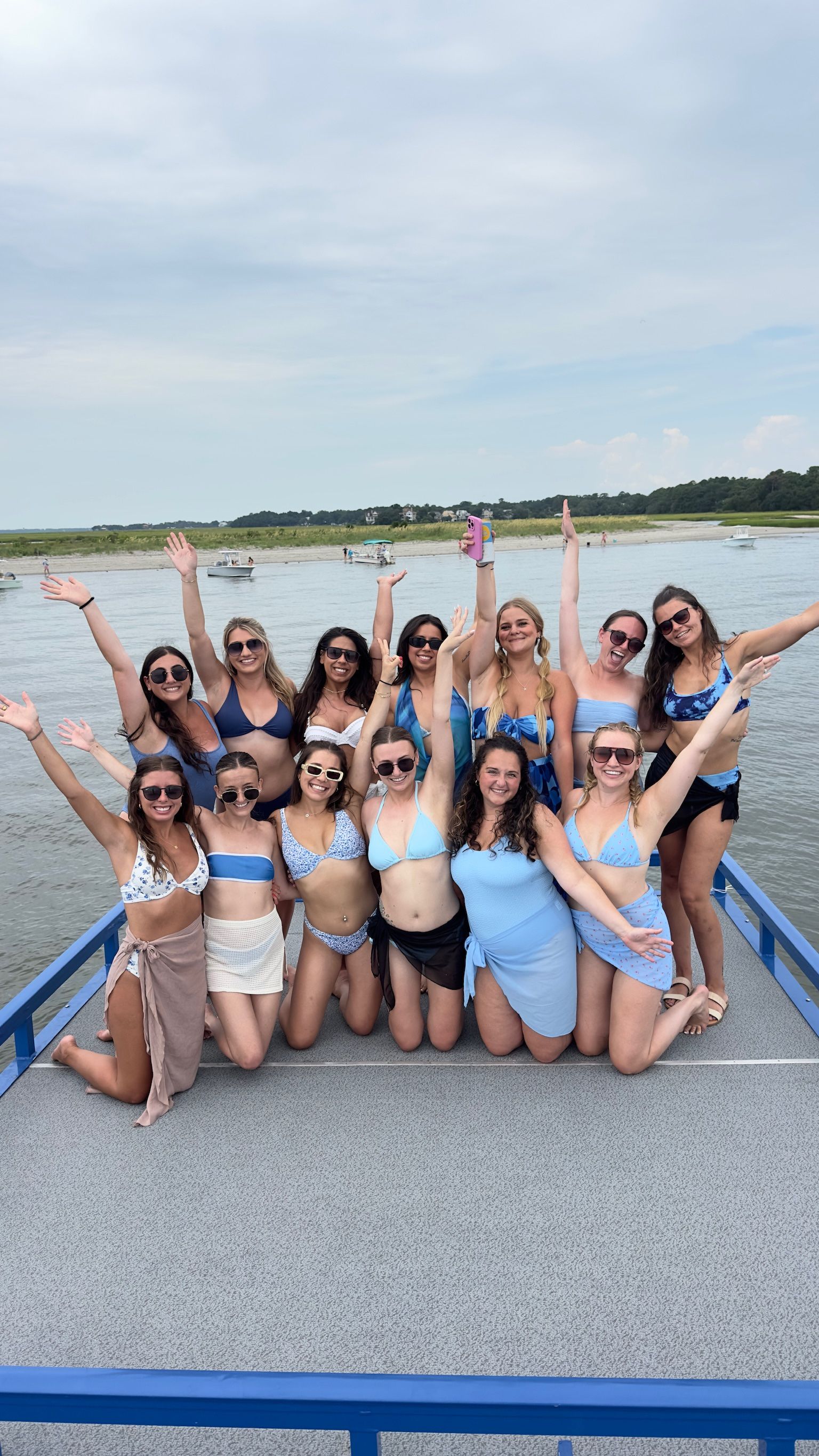 Group of friends in blue swimsuits smiling and raising their arms while posing on a boat deck over a calm coastal bay with marshy shoreline and small boats in the background on a summer beach day.