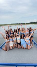Group of friends in blue swimsuits smiling and raising their arms while posing on a boat deck over a calm coastal bay with marshy shoreline and small boats in the background on a summer beach day.