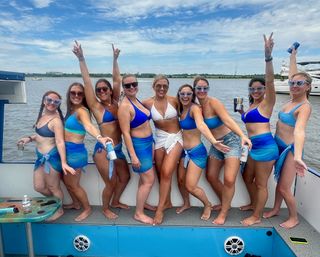 Group of friends in blue bikinis and matching sarongs, plus one in white, wearing sunglasses and holding drinks while posing and cheering on a summer boat outing at a waterfront.