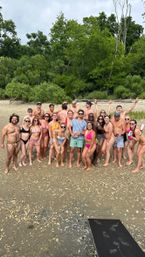 Large group of friends in colorful swimsuits smiling and posing for a beach group photo on a shell-strewn sandy shoreline with green, tree-lined background on a cloudy summer day.