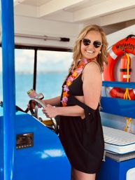 Smiling woman in heart-shaped sunglasses and a colorful lei steering a bright blue boat on clear tropical ocean waters with a life preserver visible in the background