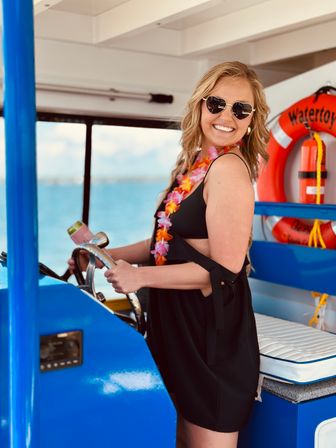 Smiling woman in heart-shaped sunglasses and a colorful lei steering a bright blue boat on clear tropical ocean waters with a life preserver visible in the background