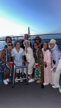 Group of friends in playful retro outfits posing on a boat at dusk with inflatable walkers and red cups, calm water and a lit cable-stayed bridge in the background.