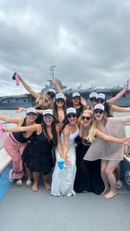 Ten women in summer outfits and matching white caps cheer on a party boat, holding colorful cups and tumblers, with a gray naval aircraft carrier and cloudy sky in the harbor behind them.