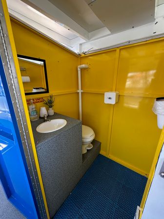 Cheery yellow portable restroom interior with a sink and faucet, small mirror, stepped toilet, wall dispenser and blue anti-slip floor