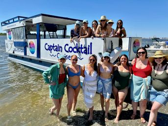 Smiling group of women in swimsuits posing on a sandy shoreline next to a colorful cocktail cruise boat on a sunny coastal bay.