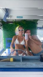 Playful boat portrait: two smiling people on a sunny boat — person in a white bikini and sunglasses at the wheel, companion holding a drink can and a cardboard smile, faux boxwood backdrop with neon décor and ocean wake outside.
