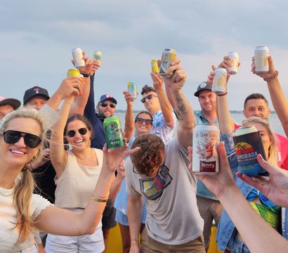 Group of friends on a boat by a calm lake, smiling and raising canned drinks in a sunset cheers.