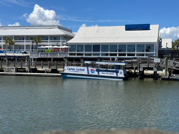 Tour boat moored at a lively waterfront restaurant and wooden marina dock on a sunny coastal harbor with blue skies