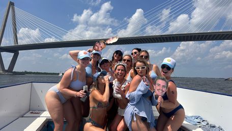 Group of women in swimsuits celebrating on a sunny boat cruise beneath a cable-stayed bridge, laughing and holding drinks and playful face cutouts with blue sky and water in the background.