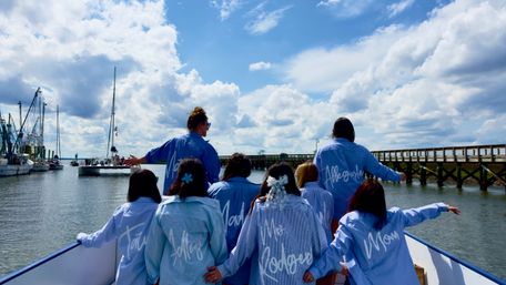 Cheerful bridal party in matching blue shirts on the bow of a boat, facing a marina with sailboats and a wooden pier under a bright, cloud-filled sky