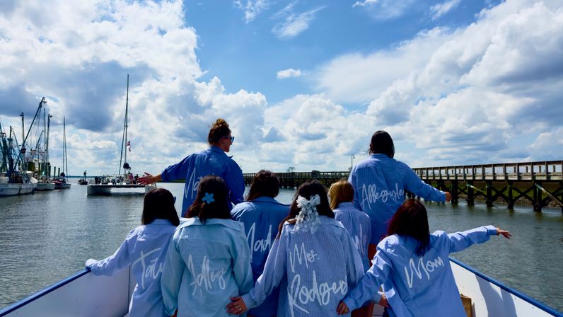 Cheerful bridal party in matching blue shirts on the bow of a boat, facing a marina with sailboats and a wooden pier under a bright, cloud-filled sky