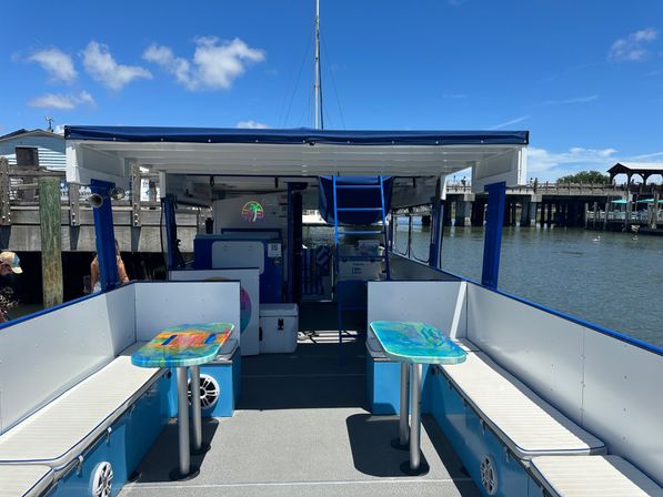 Sunny marina scene: open-deck pontoon boat with blue trim and canopy, cushioned bench seating and two colorful resin tables, dock and waterfront in the background under a bright blue sky.