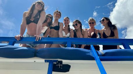 Seven women in bikinis leaning over a bright blue boat railing, smiling and enjoying a sunny, carefree day on the water