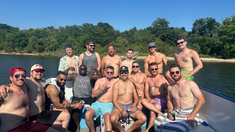 About 15 friends smiling and relaxing shirtless on a pontoon boat on a sunny lake with a wooded shoreline and clear blue sky in the background.