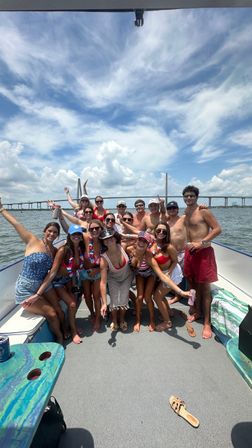 Group of friends in swimsuits enjoying a sunny boat party, posing on the deck with water and a cable-stayed bridge spanning the bay under a bright blue sky.