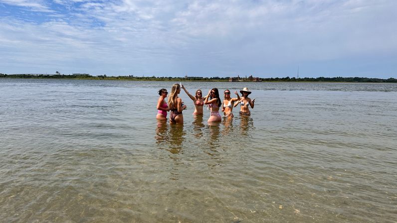 Group of six people in colorful bikinis standing waist-deep in shallow coastal water, toasting and posing near a tree-lined shoreline under a partly cloudy summer sky.