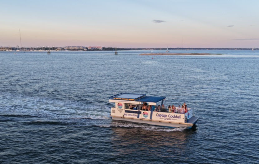 Sightseeing party boat with passengers cruising across a calm coastal bay at sunset, leaving a white wake with low shoreline and sandbar on the horizon.