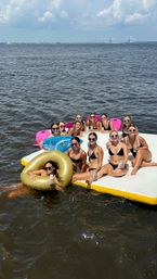Group of friends in bikinis relaxing on a floating platform and inflatables with drinks on a sunny bay, distant cable-stayed bridge and blue sky in the background