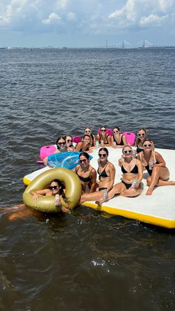 Group of friends in bikinis relaxing on a floating platform and inflatables with drinks on a sunny bay, distant cable-stayed bridge and blue sky in the background
