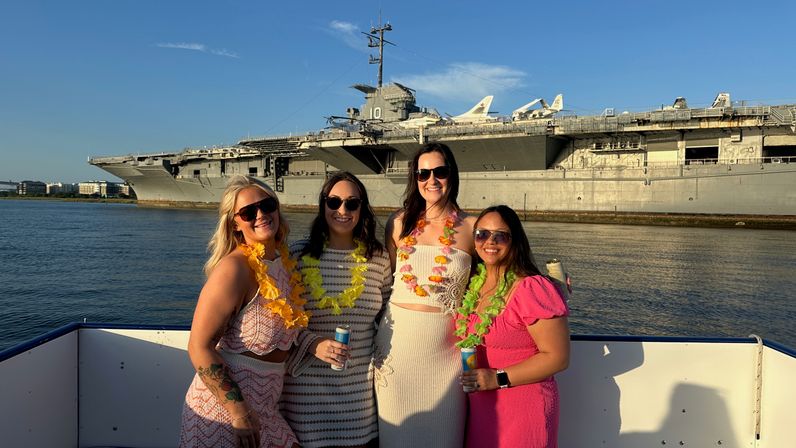 Four friends in summer dresses and sunglasses wearing colorful leis on a boat in front of a docked aircraft carrier at golden hour over a calm harbor