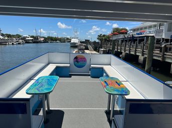 Sunny open-deck boat with white bench seating and two colorful tables, docked at a marina with boats and waterfront restaurants under a blue sky