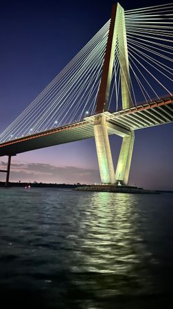 Dramatic cable-stayed bridge at twilight with illuminated concrete pylon and fan of suspension cables, green lights reflecting on rippling river water