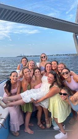 Boat party of smiling women lifting a woman in a white dress on a coastal waterway beneath a large cable-stayed bridge and blue sky