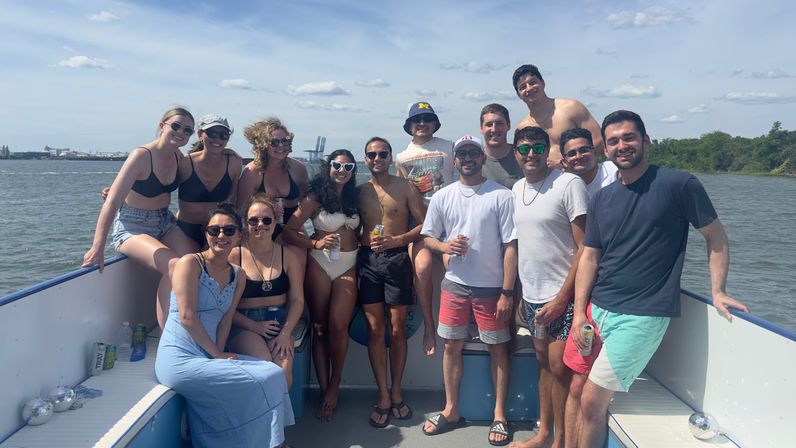 Cheerful group of young adults on a summer boat party in a coastal harbor, wearing swimsuits and shorts, holding drinks under a blue sky with distant shoreline and industrial cranes