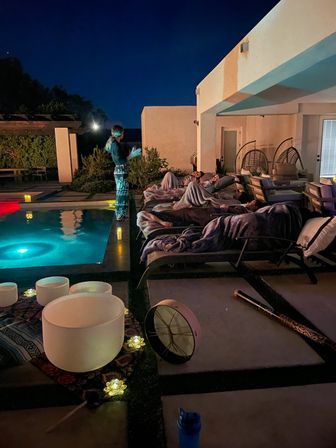 Nighttime backyard poolside sound bath on a modern patio — people wrapped in blankets on lounge chairs beside an illuminated pool, crystal singing bowls, drum and candle lights.