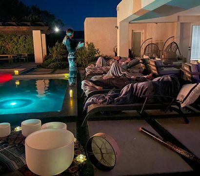 Nighttime backyard poolside sound bath on a modern patio — people wrapped in blankets on lounge chairs beside an illuminated pool, crystal singing bowls, drum and candle lights.