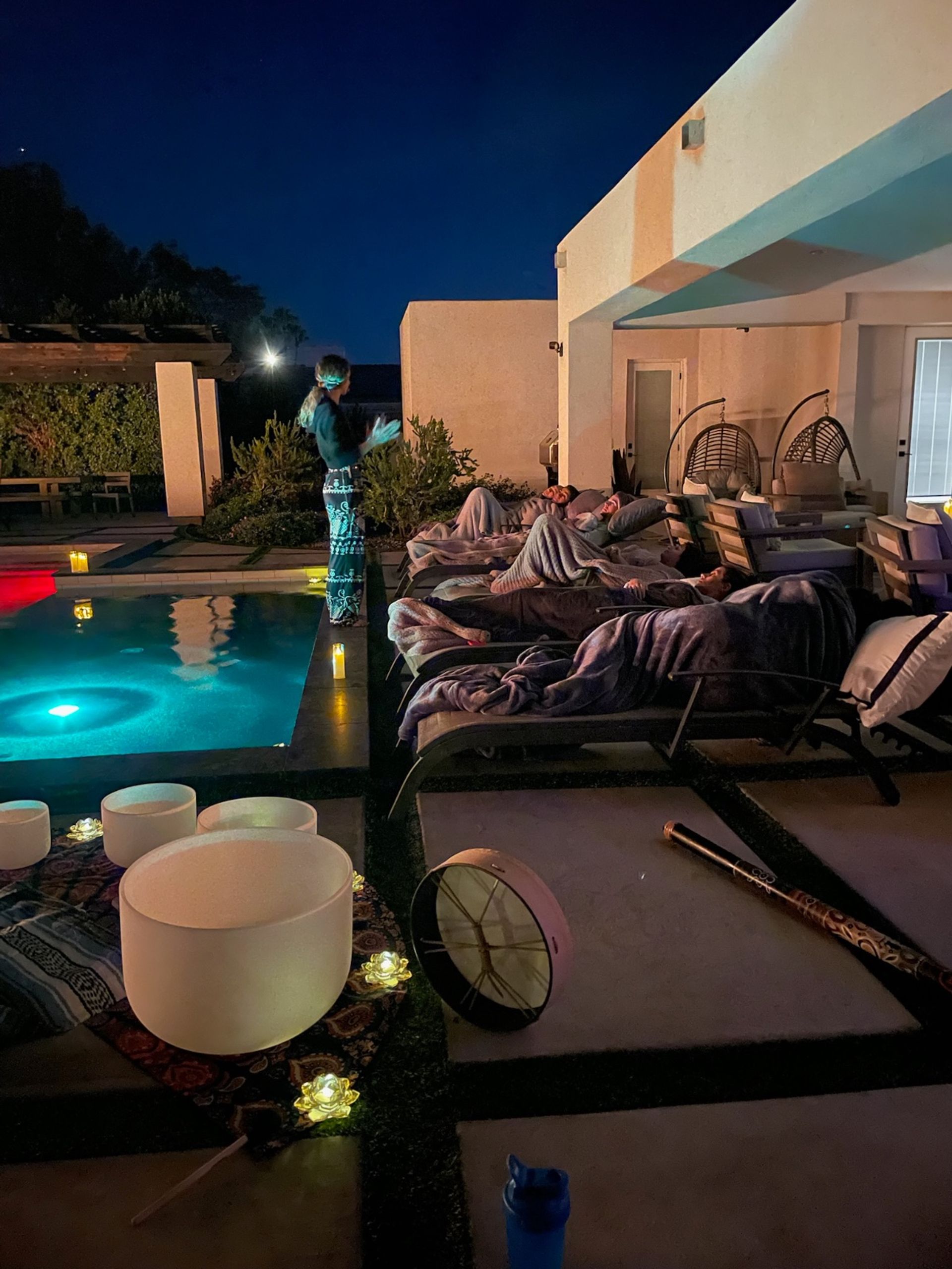 Nighttime backyard poolside sound bath on a modern patio — people wrapped in blankets on lounge chairs beside an illuminated pool, crystal singing bowls, drum and candle lights.