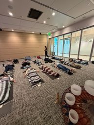 Indoor wellness scene with participants lying on mats wrapped in striped blankets in a modern conference-style room during a guided relaxation/sound bath — instructor stands near glass wall while crystal singing bowls sit in the foreground.