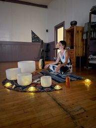 Person seated cross-legged in a meditation studio leading a sound bath with crystal singing bowls, mallets, a laptop and glowing lotus candles on a patterned rug and wooden floor.