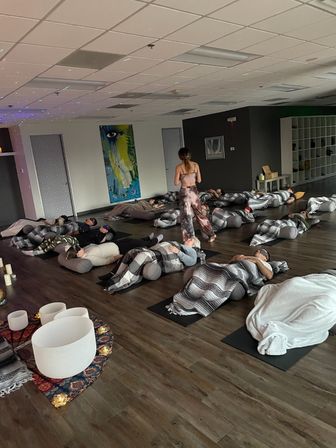 Group sound bath and guided relaxation in an indoor yoga studio — participants lying on mats wrapped in blankets with bolsters, instructor walking between rows, crystal singing bowls and candles in foreground.