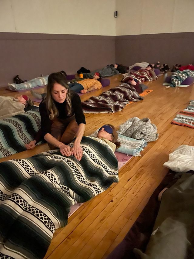 Cozy yoga studio group relaxation: rows of people lying on mats covered with patterned blankets and eye masks while a practitioner gently places hands over a participant during a guided meditation/sound bath.