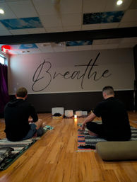 Calm indoor meditation/yoga studio with large 'Breathe' wall script, two participants seated on woven mats facing crystal singing bowls and a lit candle on a hardwood floor — sound bath setup.