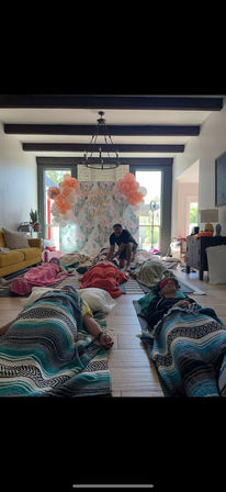 Bridal spa day in a bright living room with guests lying on mats under colorful blankets and eye masks, a therapist kneeling between rows, and a floral balloon backdrop reading a pre-wedding message.