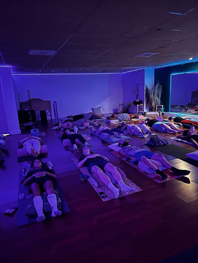 Dim indoor yoga studio with purple-blue LED lights, about a dozen people lying on mats and bolsters in Savasana for a group relaxation/sound bath session, mirror and plants visible at the front.