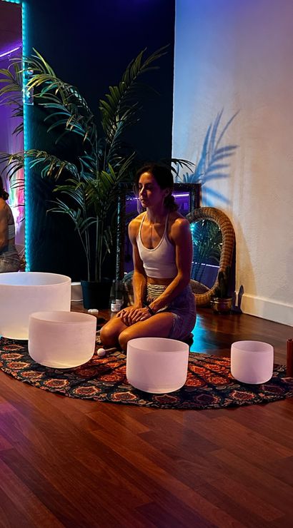 Person kneeling in a dim yoga studio during a sound bath with crystal singing bowls on a patterned boho rug, indoor palm, mirror and colorful LED mood lighting.