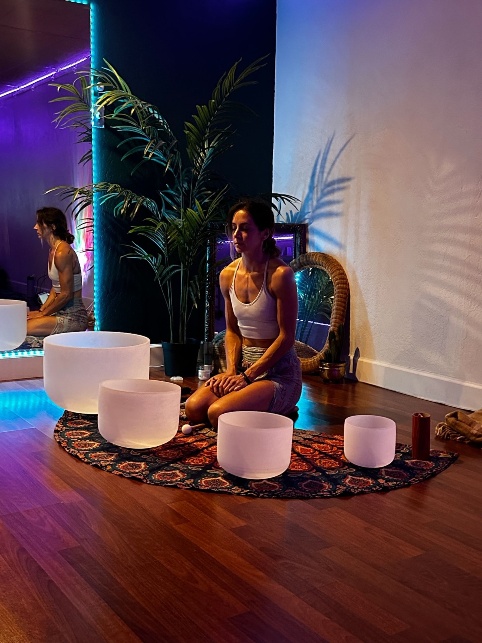 Person kneeling in a dim yoga studio during a sound bath with crystal singing bowls on a patterned boho rug, indoor palm, mirror and colorful LED mood lighting.