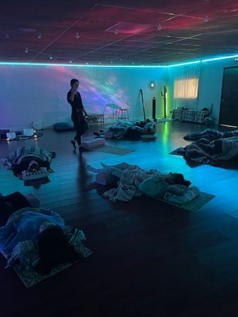 Dim wellness studio with teal and purple LED lighting, people lying on mats and blankets during a guided sound bath meditation while an instructor walks between rows.