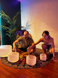 Two adults seated in a cozy indoor meditation space playing crystal singing bowls on a patterned round rug, colorful ambient lighting and a potted palm casting shadows — sound bath wellness session.