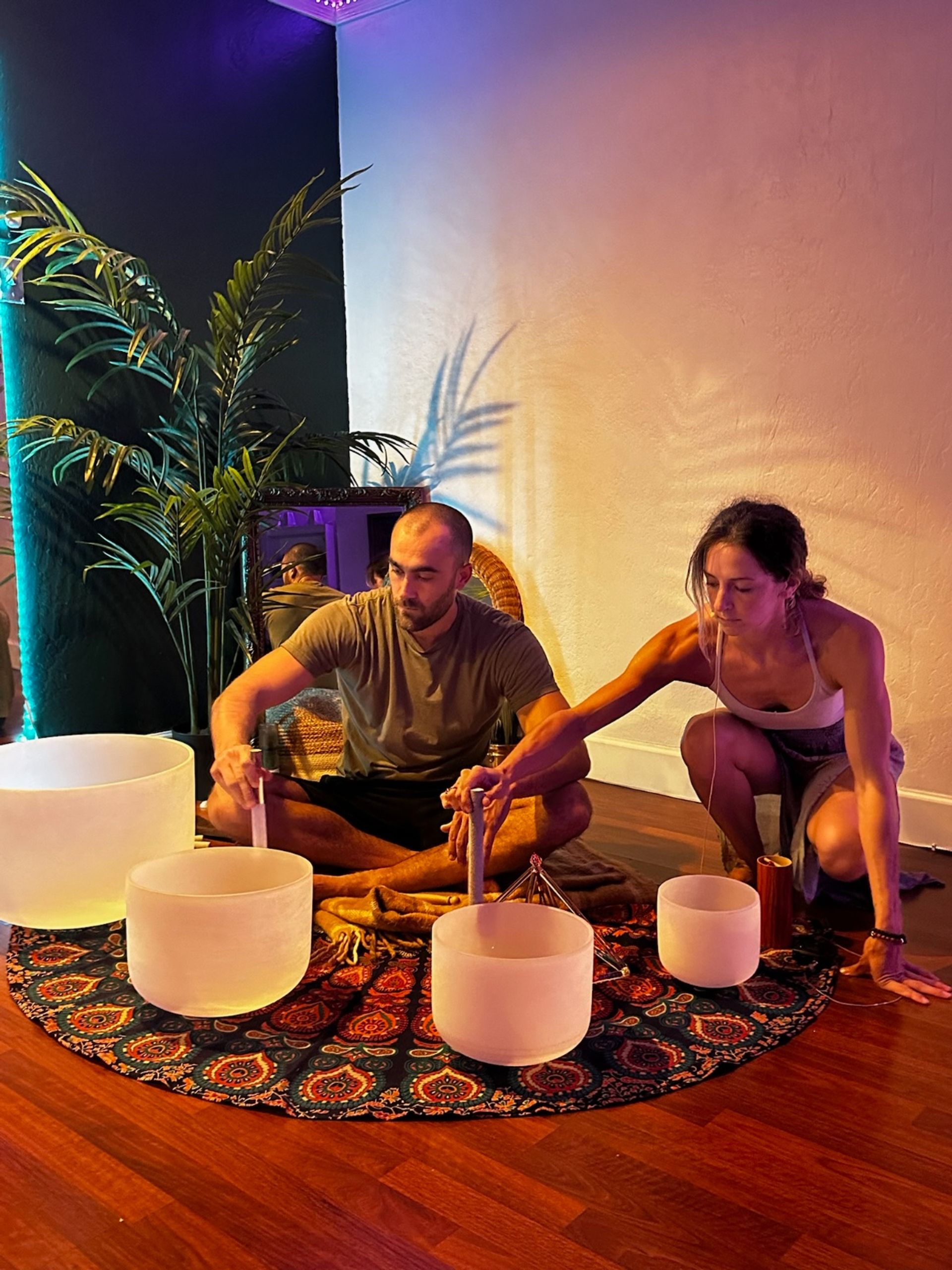 Two adults seated in a cozy indoor meditation space playing crystal singing bowls on a patterned round rug, colorful ambient lighting and a potted palm casting shadows — sound bath wellness session.