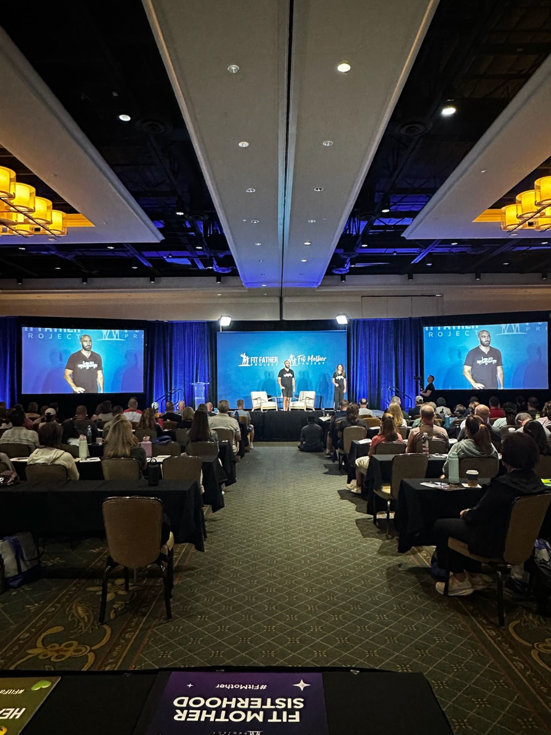 Wide hotel ballroom conference scene with a packed audience facing a central stage where two presenters speak under blue lighting, flanked by large projection screens, rows of tables and chairs, and overhead chandeliers — lively health and fitness seminar setting.