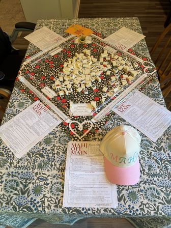 Overhead view of a ready-to-play mahjong spread on a floral dining table: scattered ivory tiles in the center, four clear clear tile racks, printed rulesheets at each seat, and a pastel pink-and-yellow cap embroidered with 'MAHJ'.