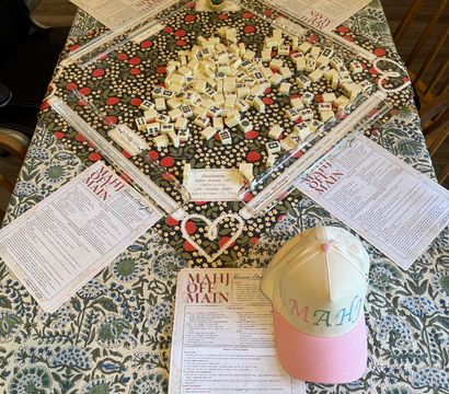 Overhead view of a ready-to-play mahjong spread on a floral dining table: scattered ivory tiles in the center, four clear clear tile racks, printed rulesheets at each seat, and a pastel pink-and-yellow cap embroidered with 'MAHJ'.