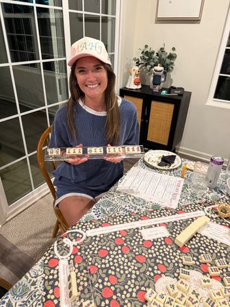 Smiling person wearing a pastel cap holding a mahjong tile rack at a cozy home dining table, surrounded by mahjong tiles, a score sheet, snacks and drinks on a patterned tablecloth.