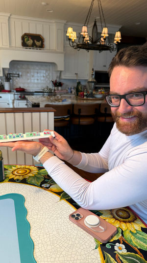 Bearded man holding a rack of pastel mahjong tiles at a bright farmhouse-style kitchen island with sunflower tablecloth and chandelier, game-night vibe.