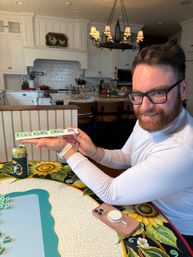 Smiling bearded man in glasses holding a mahjong tile rack at a sunflower-print dining table in a bright white farmhouse-style kitchen with pendant chandelier